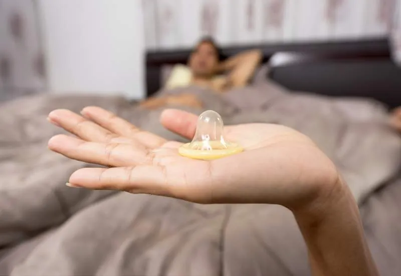 Hand hält ein Kondom, im Hintergrund liegt eine Person im Bett